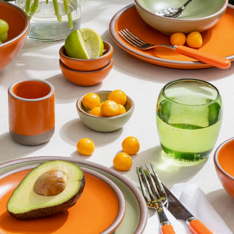 On a table is ceramic plates, bowls, and cups in bold orange and soft sage green colors, a green tumbler glass filled with water, orange-handled fork and knife, and little tomatoes and a sliced avocado.