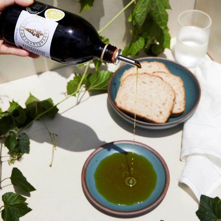 A hand holding a dark bottle of olive oil that is pouring onto plate next to a bowl of sliced bread