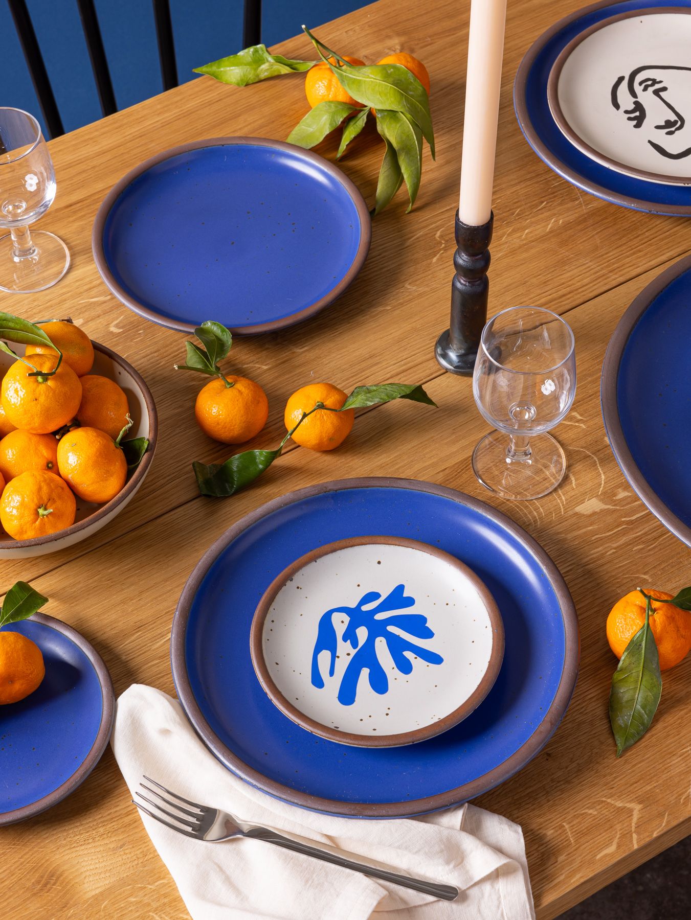 A wooden dining table set with vibrant blue La Sirène plates from East Fork’s Matisse Collection. A small plate with a blue decal of a cut-paper-inspired leaf sits atop a larger blue plate. Nearby, there are clementines with leaves still attached, a tall candle, water glasses, and neatly folded napkins, creating a lively, artful tablescape.