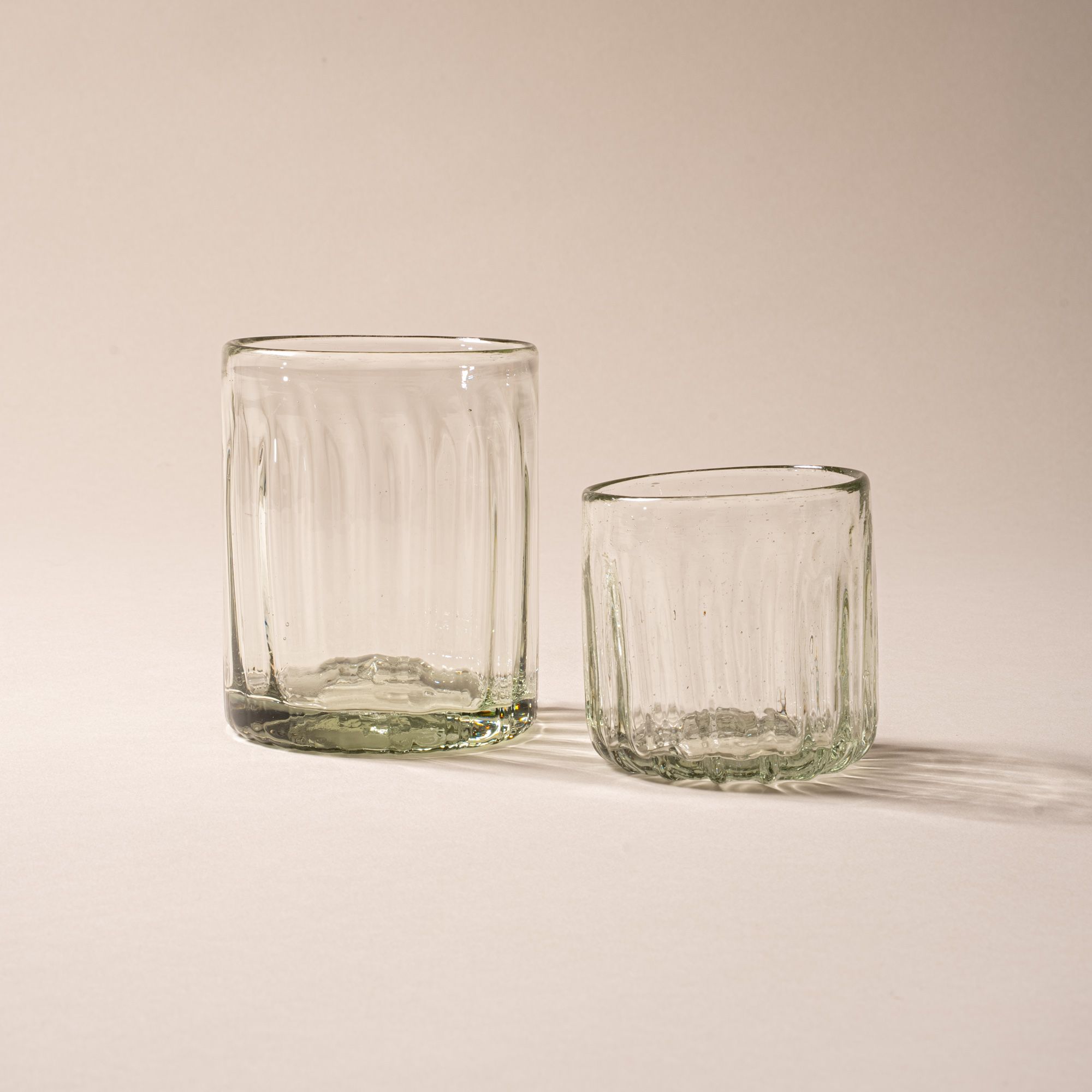 Two clear glass tumblers with vertical ridges, one tall and one short, on a neutral backdrop.