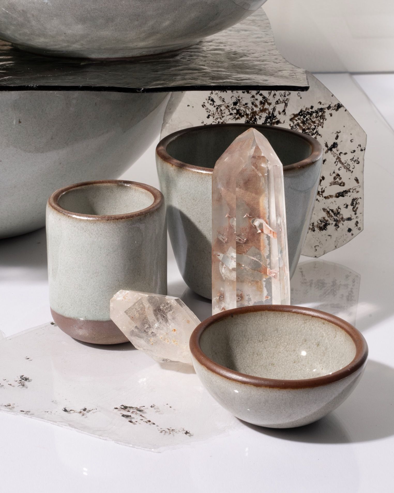 Reflective icy white ceramic cups and bowls with unglazed rims arranged beside clear quartz crystals on a reflective white surface.