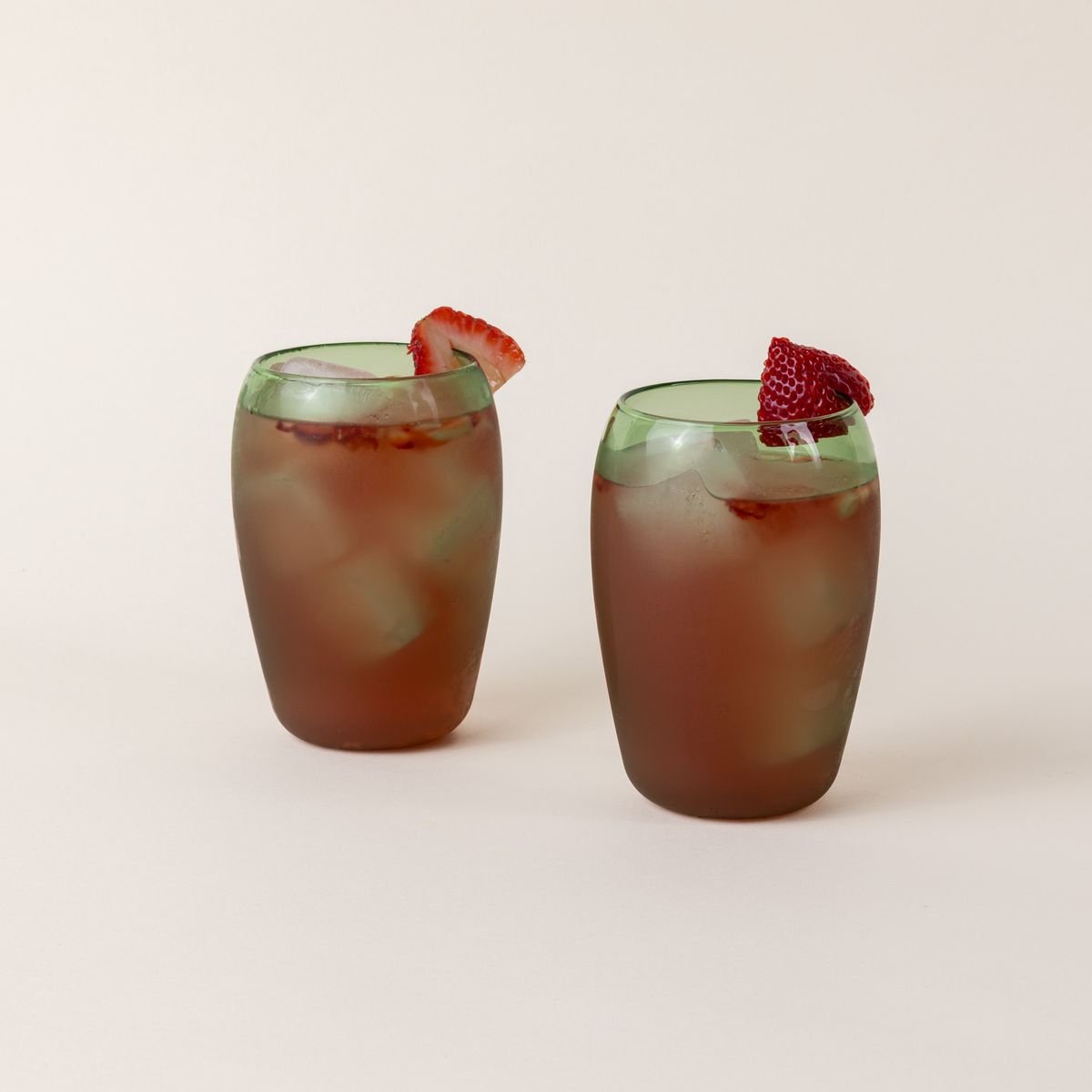 Two tall light green glass tumblers with a slight curve inward on the top. The glasses are filled with a red cocktail with sliced strawberries on the rim.