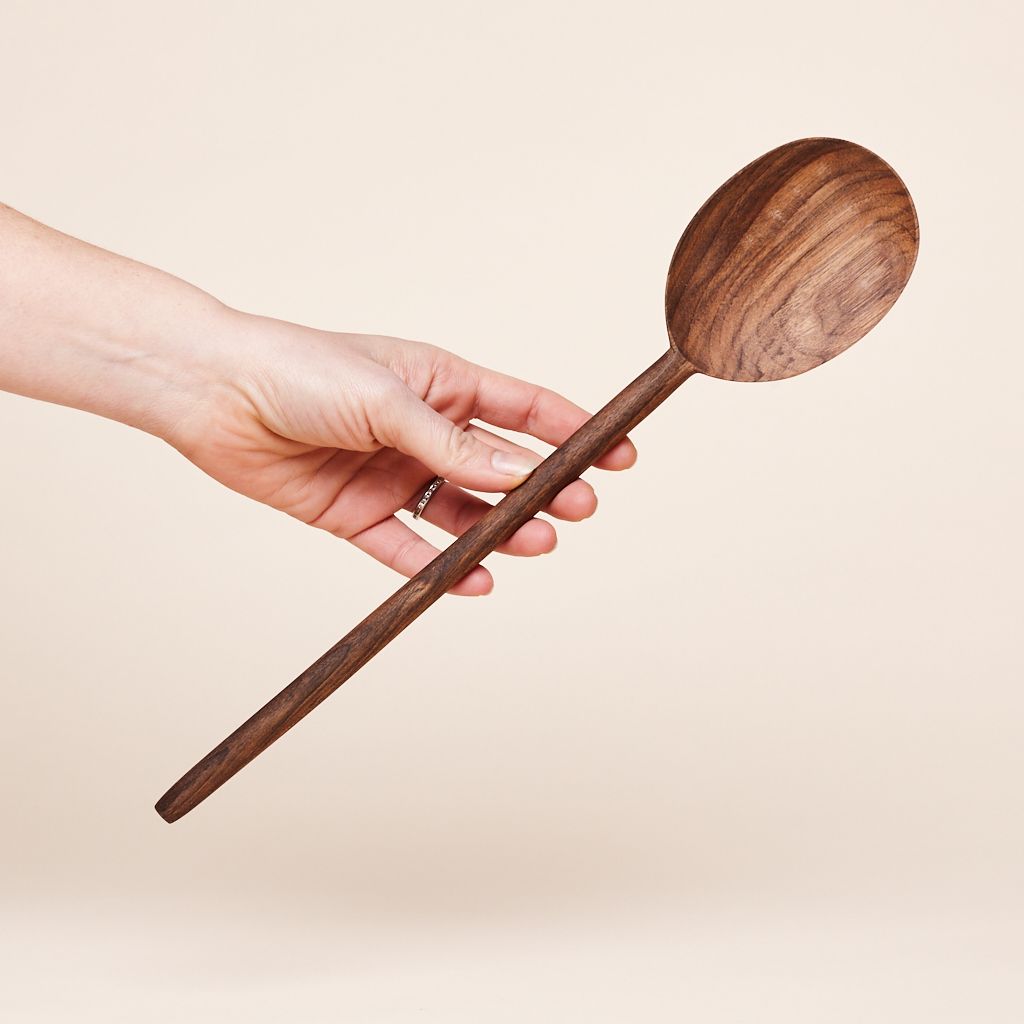 A hand with fingers and thumb extended, holding the long, thin handle of a dark brown wooden spoon