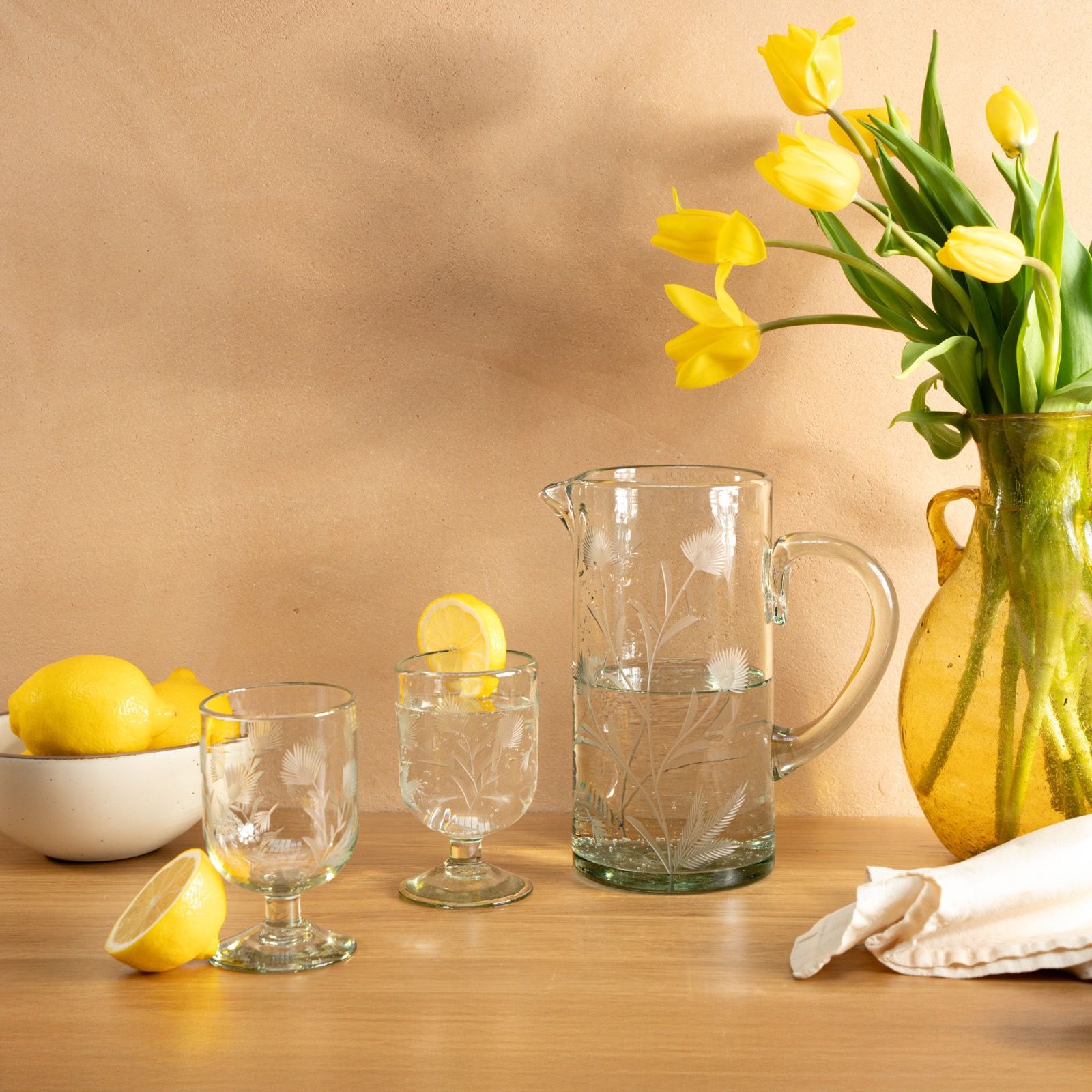 Etched thistle wine glasses and pitcher sit on a wooden table next to a bowl of lemons and vase of tulips.