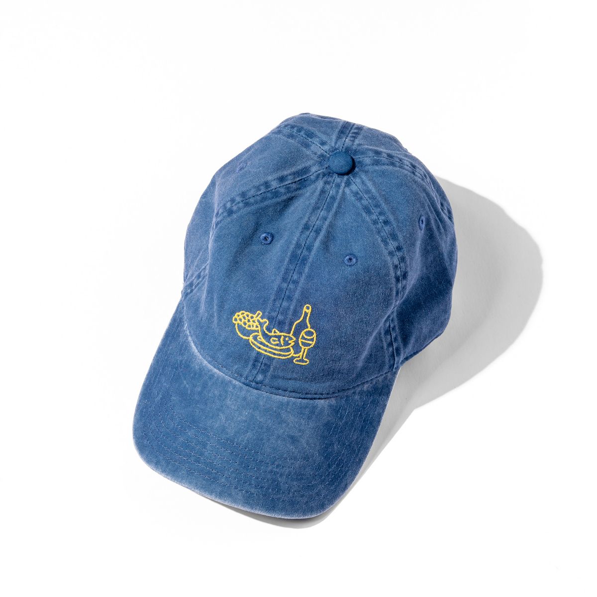 A classic blue baseball hat with yellow embroidery on the front depicting a feast of fish, fruit and wine.