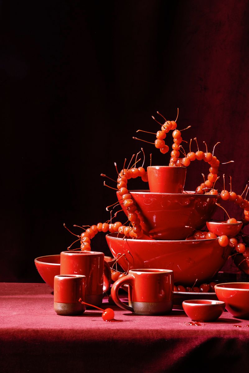 Glossy red ceramic cups, mugs and bowls stacked into a tower, draped with glossy maraschino cherries against a deep red backdrop.