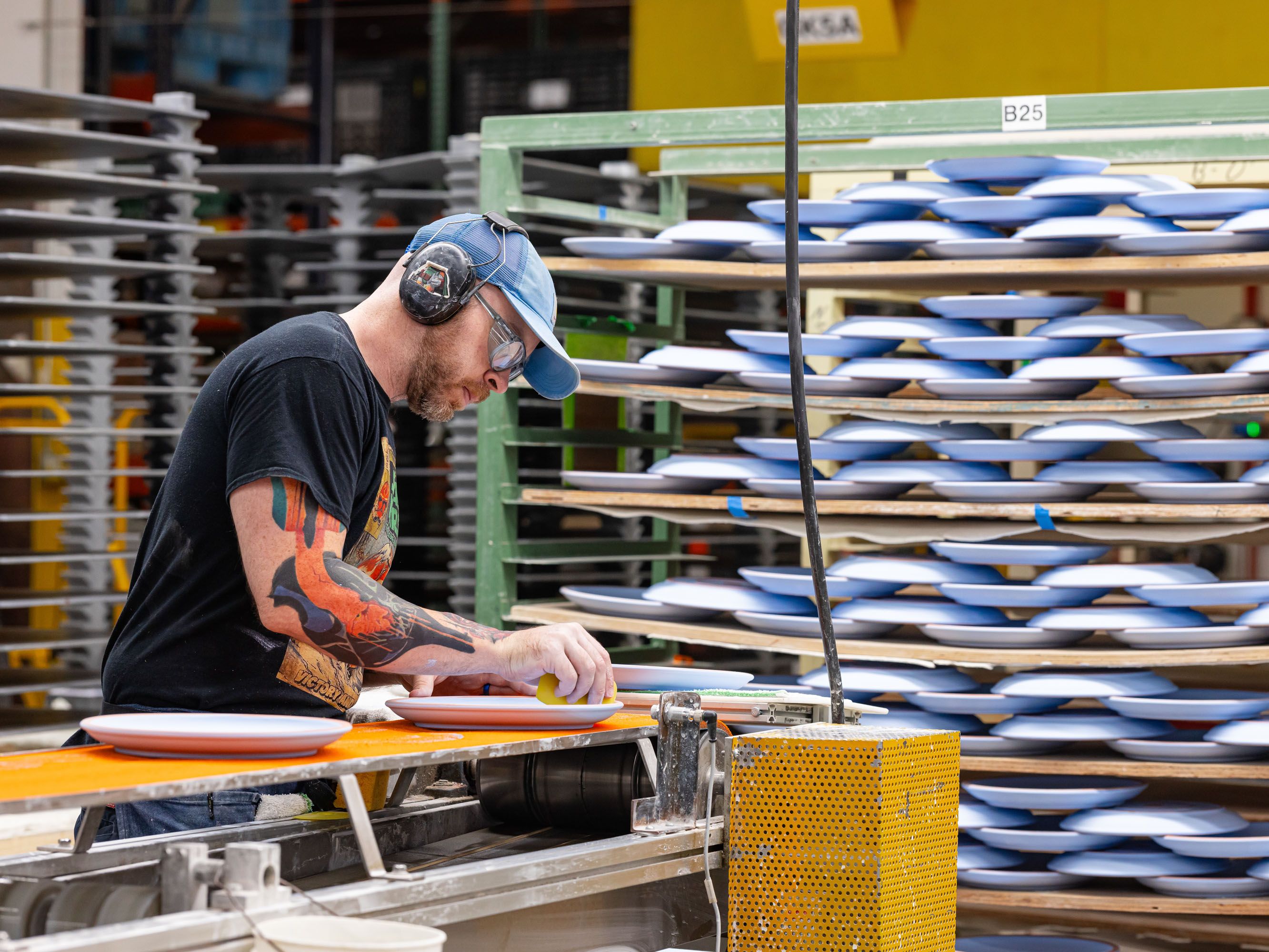 In a factory setting, a person is working on ceramic plates and there are large shelves holding periwinkle ceramic plates