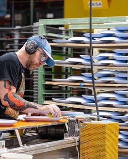 In a factory setting, a person is working on ceramic plates and there are large shelves holding periwinkle ceramic plates