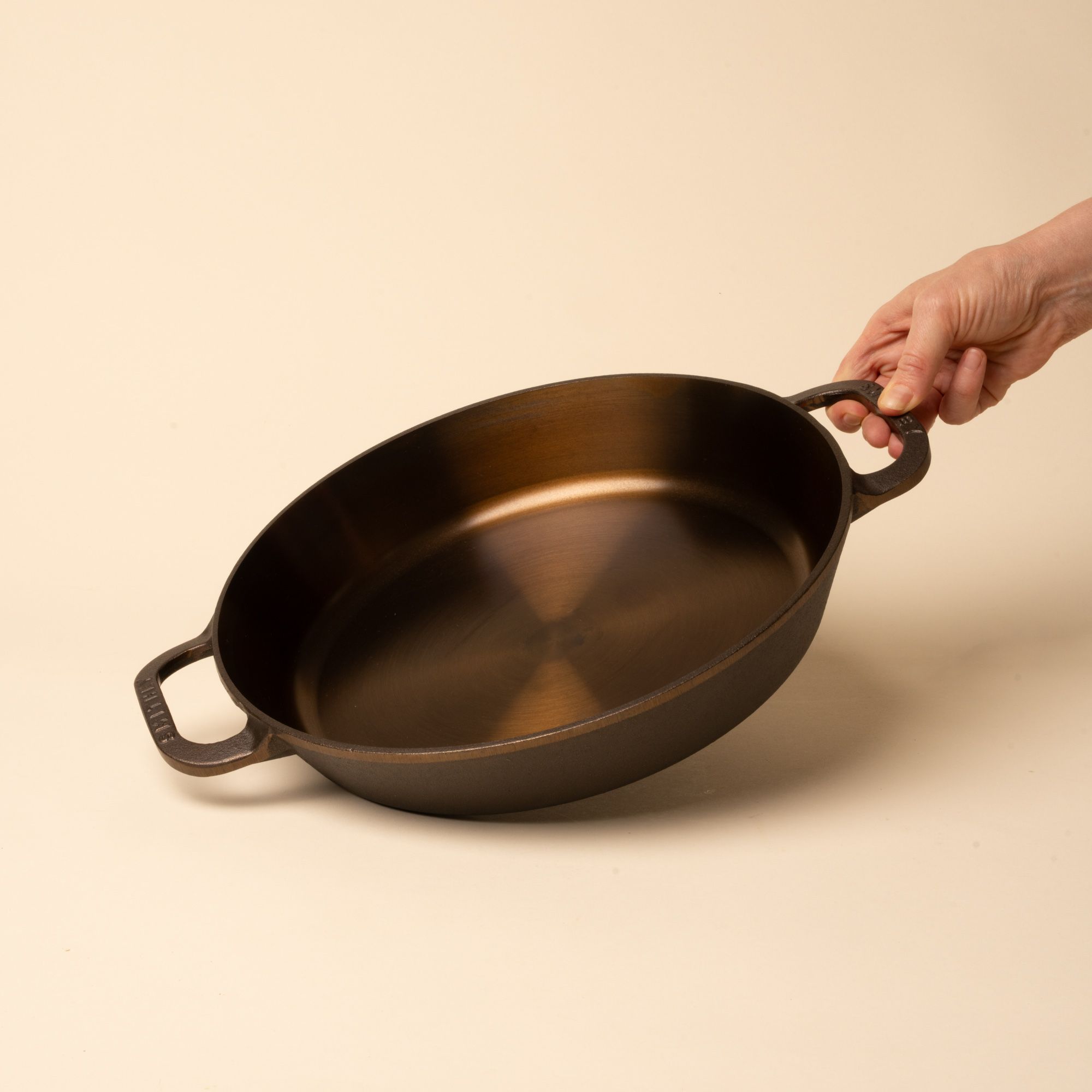 Hand holds a round cast iron pan with two side handles on a beige background.