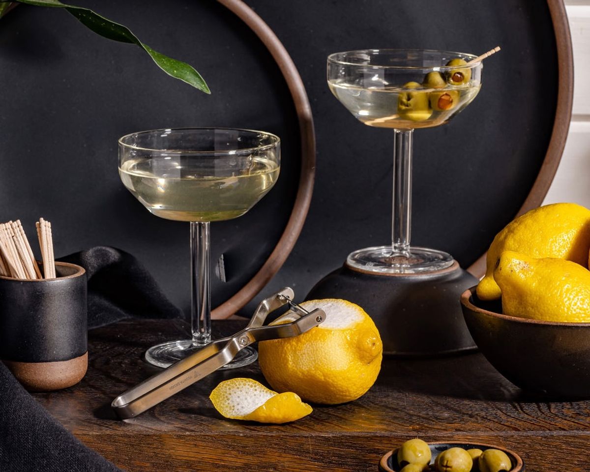 Two clear martini glasses filled with martinis. Nearby is a bowl of lemons, a lemon with a stainless steel peeler, and a little bowl of olives.