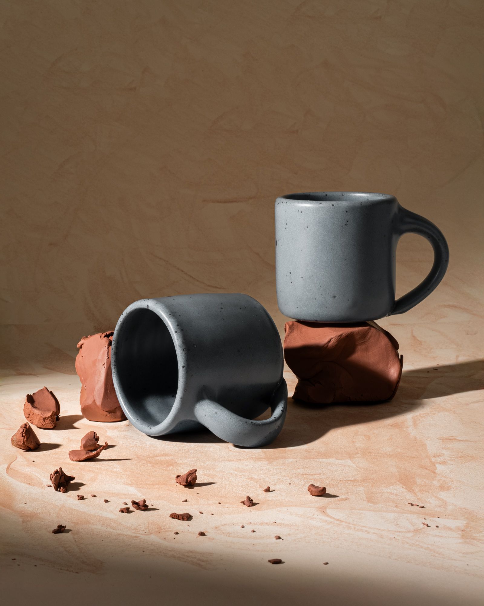 Two medium sized ceramic mugs with handles in a light blue-grey color featuring iron speckles, styled with raw clay