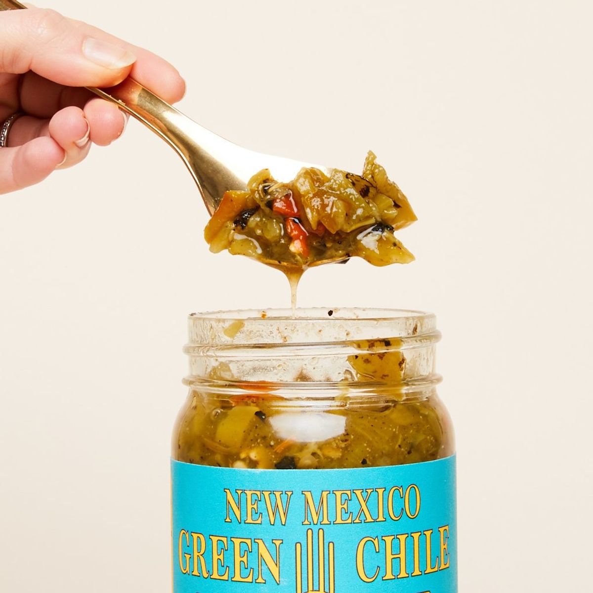 A spoon full of green salsa is held by a hand above a jar of the same salsa. Label reads, "New Mexico Green Chile"