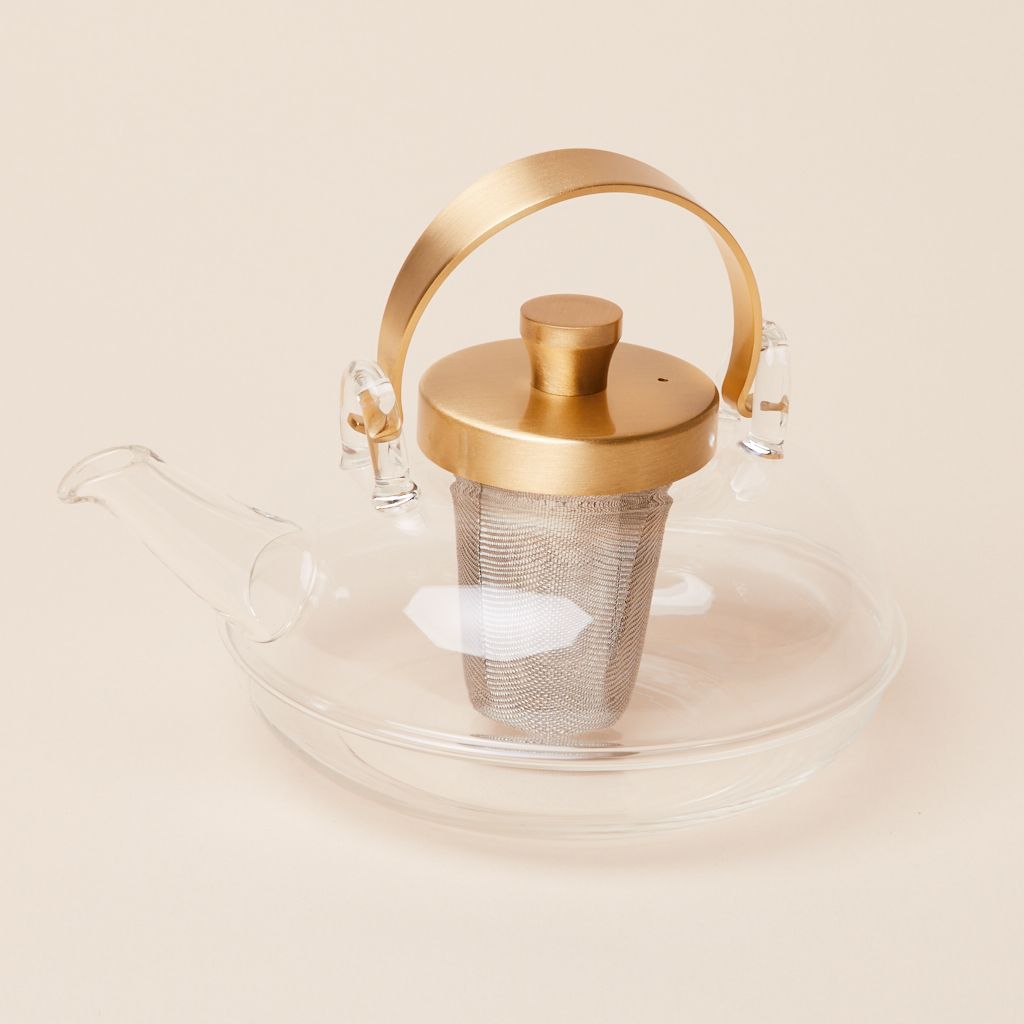 Tea pot made of glass with a glass spout with a gold handle, gold lid and a gray metal silver insert for loose tea