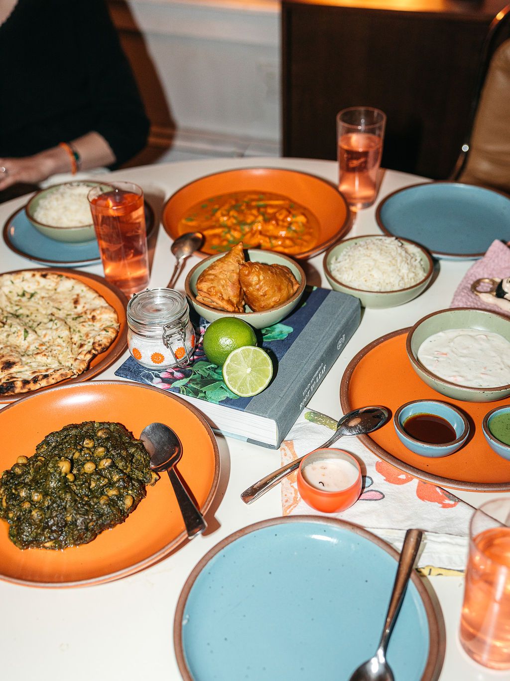 A dinner party table filled with various ceramic plates and bowls in bold colors, drinking glasses, and glimpses of people.