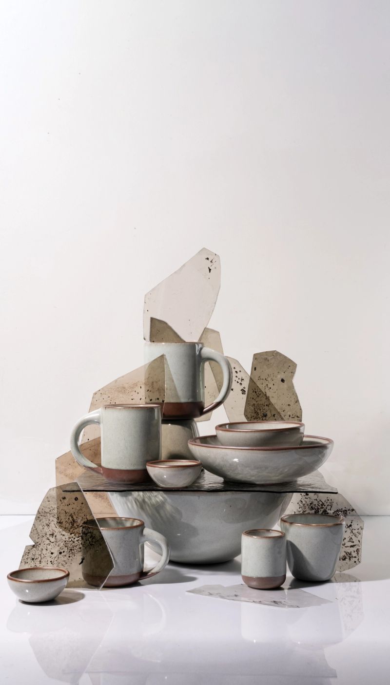 Arrangement of reflective icy white ceramic mugs and bowls with unglazed rims, displayed with textured glass panels against a light background.