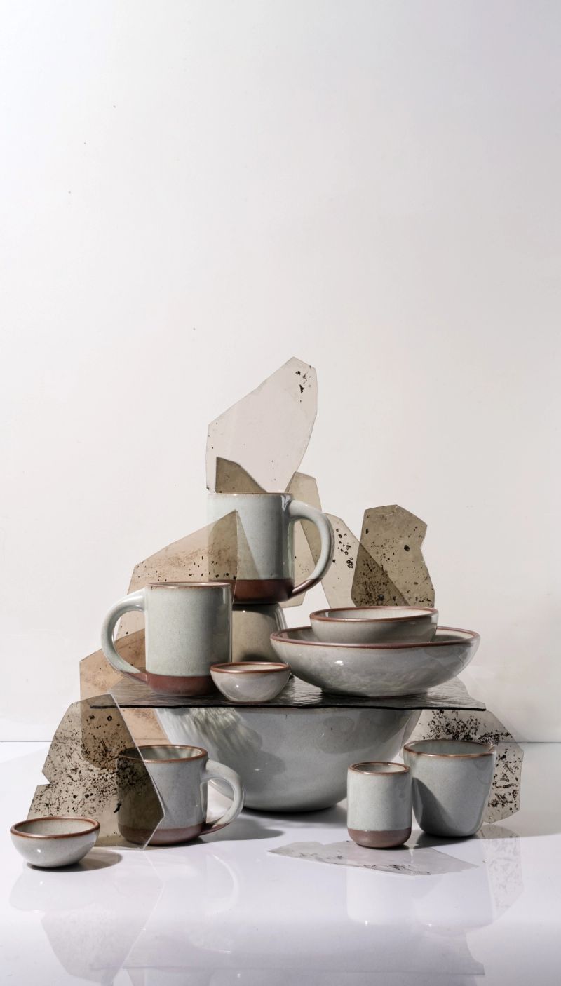 Arrangement of reflective icy white ceramic mugs and bowls with unglazed rims, displayed with textured glass panels against a light background.