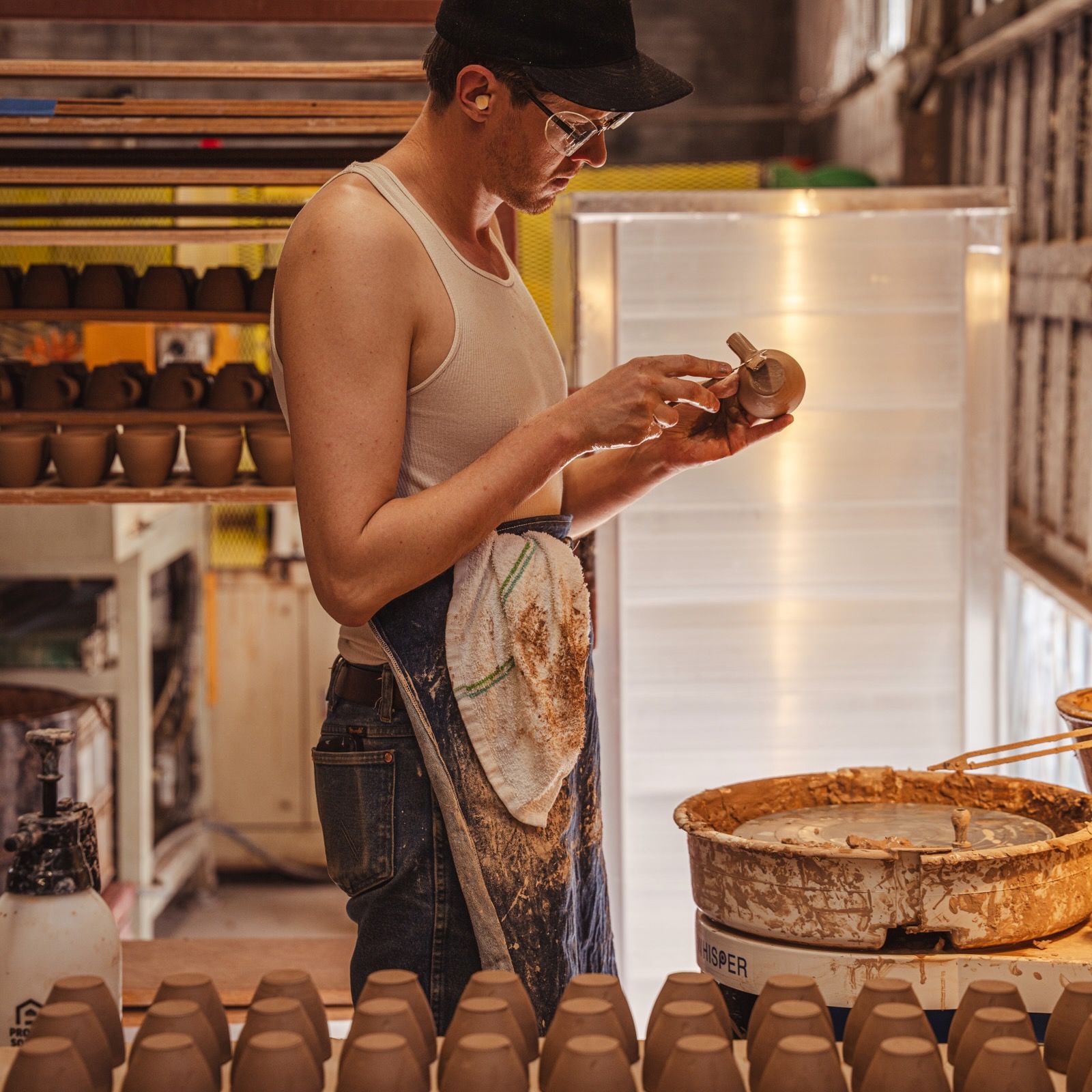 John adding a handle on an espresso cup next to a potter's wheel and in front of a table filled with espresso cups in progress.
