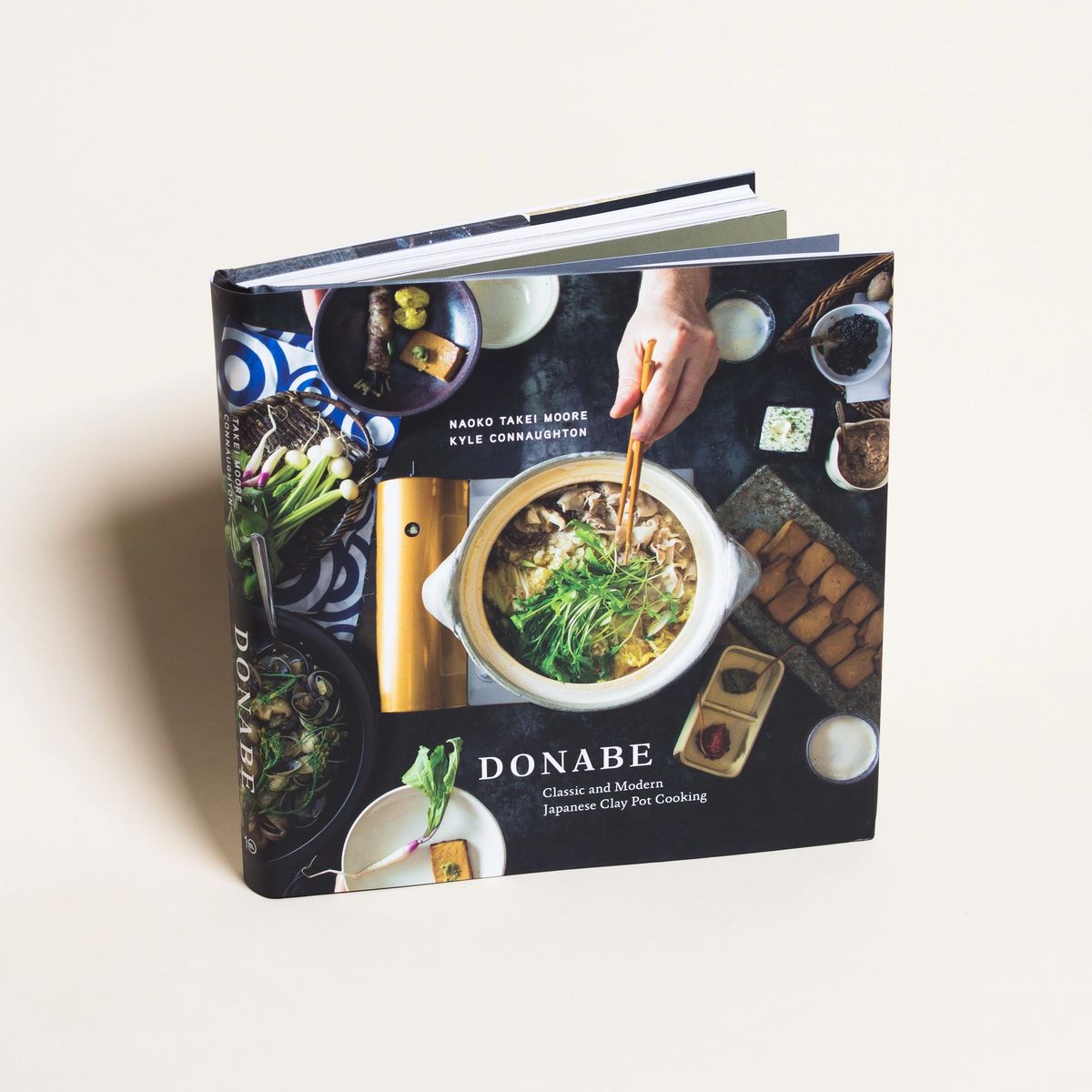 A book shown at a slight angle and the word "Donabe," and a clay pot filled with soup and greens surrounded by bowls and ingredients, a hand reaching in with chopsticks