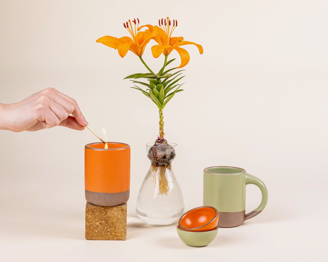A hand lights a candle in a ceramic orange vessel. Nearby is a glass bulb vase, a sage green ceramic mug, and 2 tiny bowls in bold orange and sage green colors.