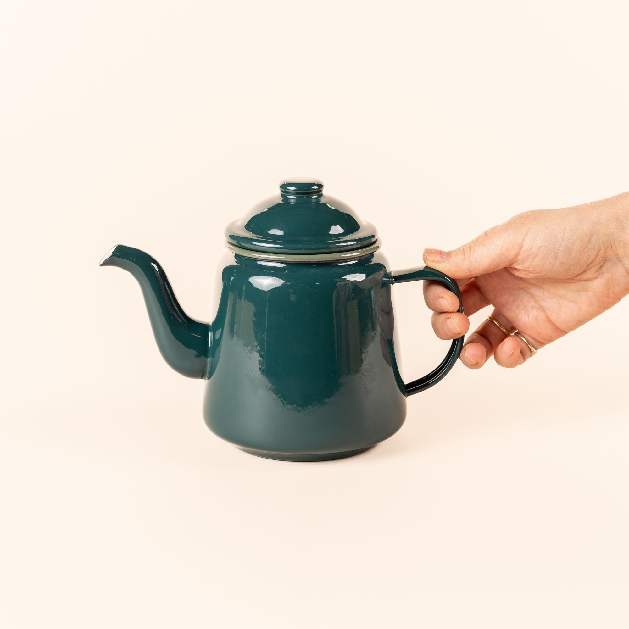 A hand holds the thin handle of a deep dark teal enamel teapot with a large spout, and simple lid