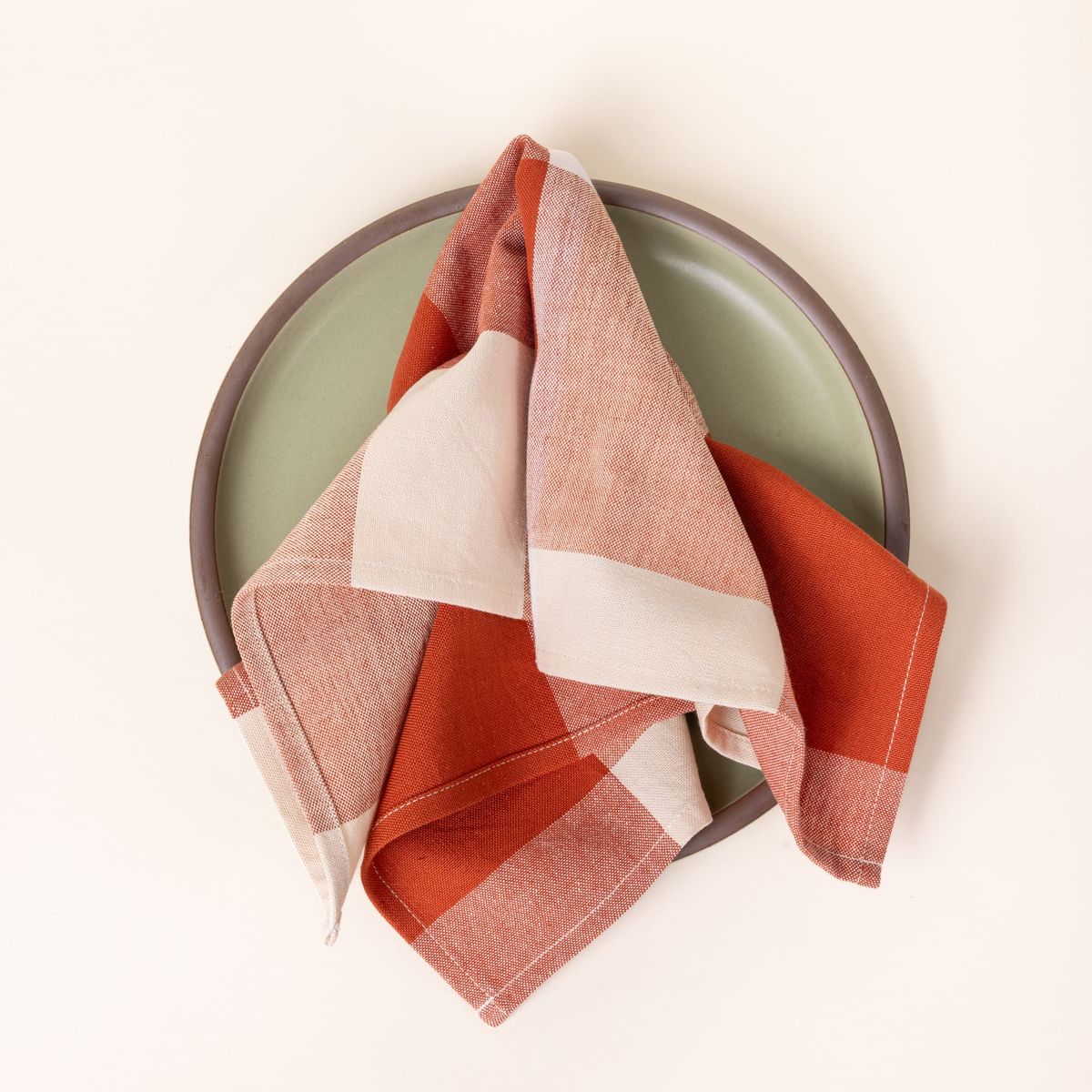 A square dinner napkin in a rust and cream gingham pattern displayed on a calming sage green ceramic dinner plate