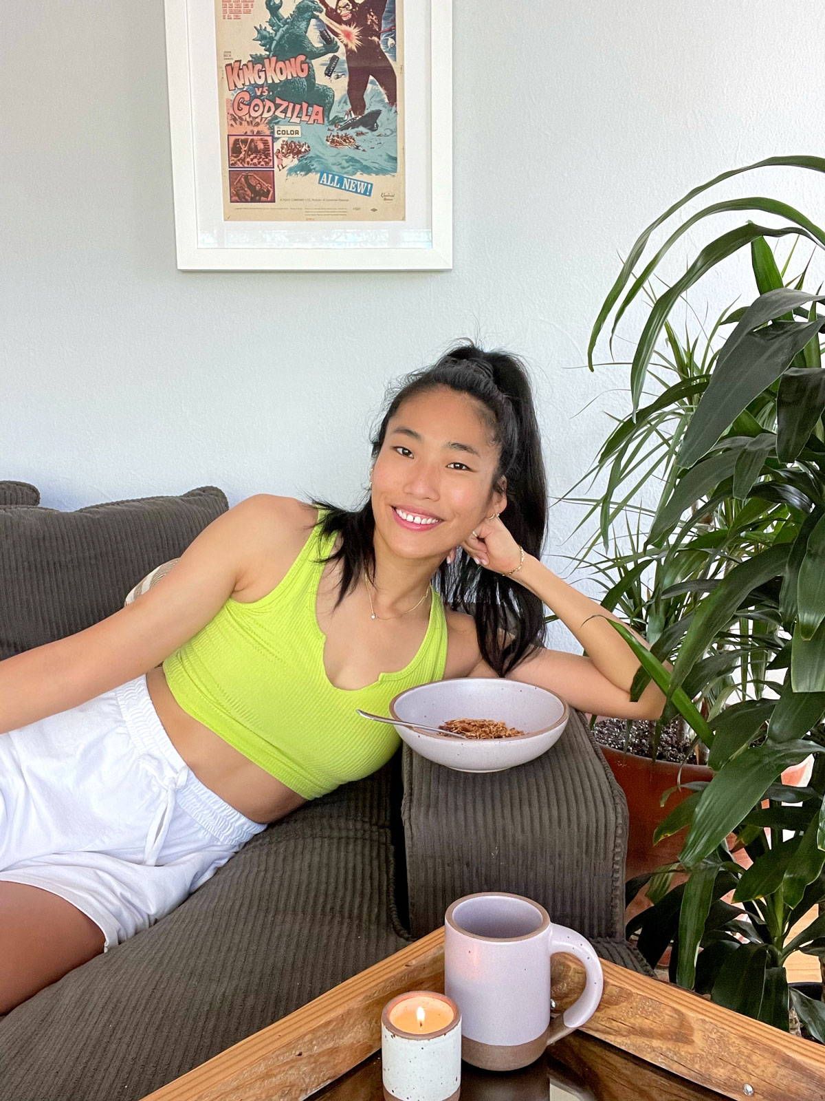 Vera is sitting on a couch and smiling next to a large plant, and ceramic bowl, mug, and candle.