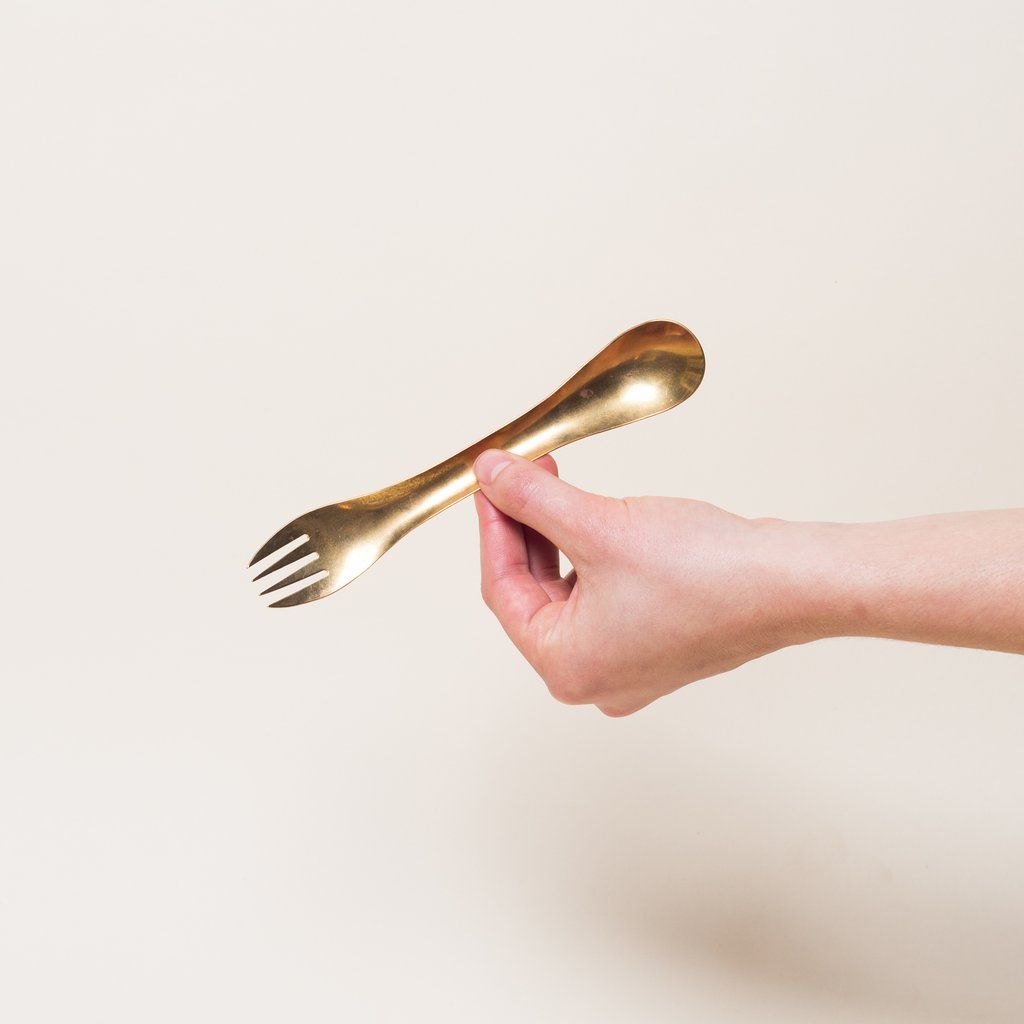 A hand holds a brass eating utensil with spoon at one end and fork at the other