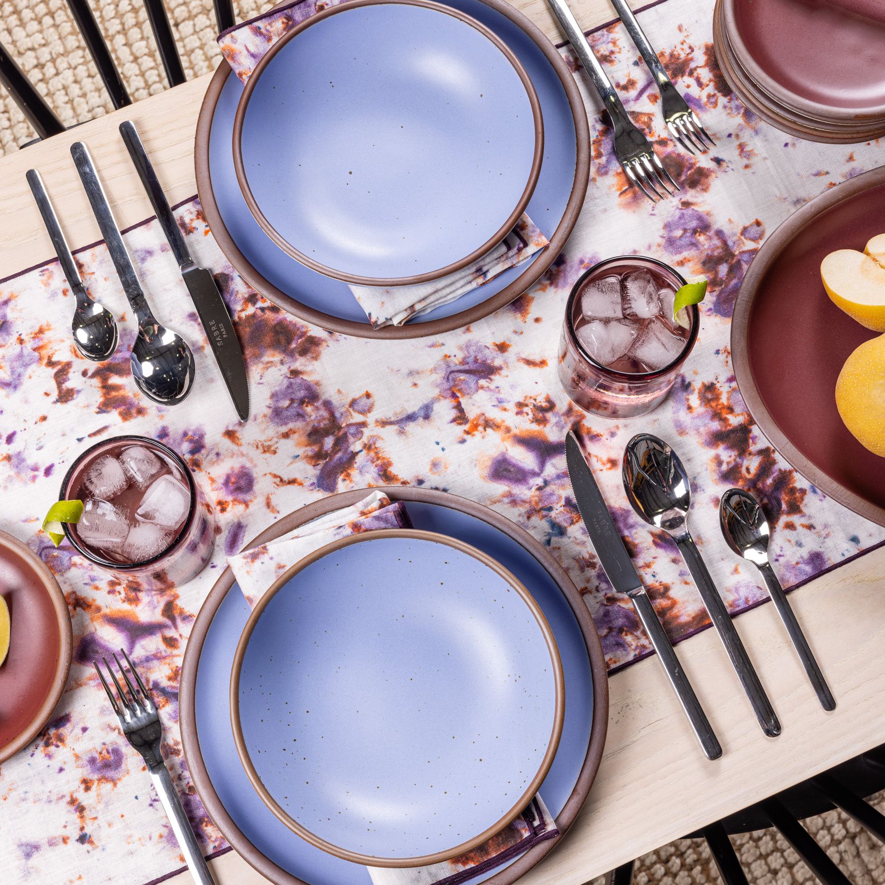 Two place settings on a marbled purple and white tablecloth with subtle hints of orange. Each setting is a periwinkle ceramic dinner plate, bowl, and napkin matching the tablecloth. The settings are surrounded by modern flatware and colorful glassware. 