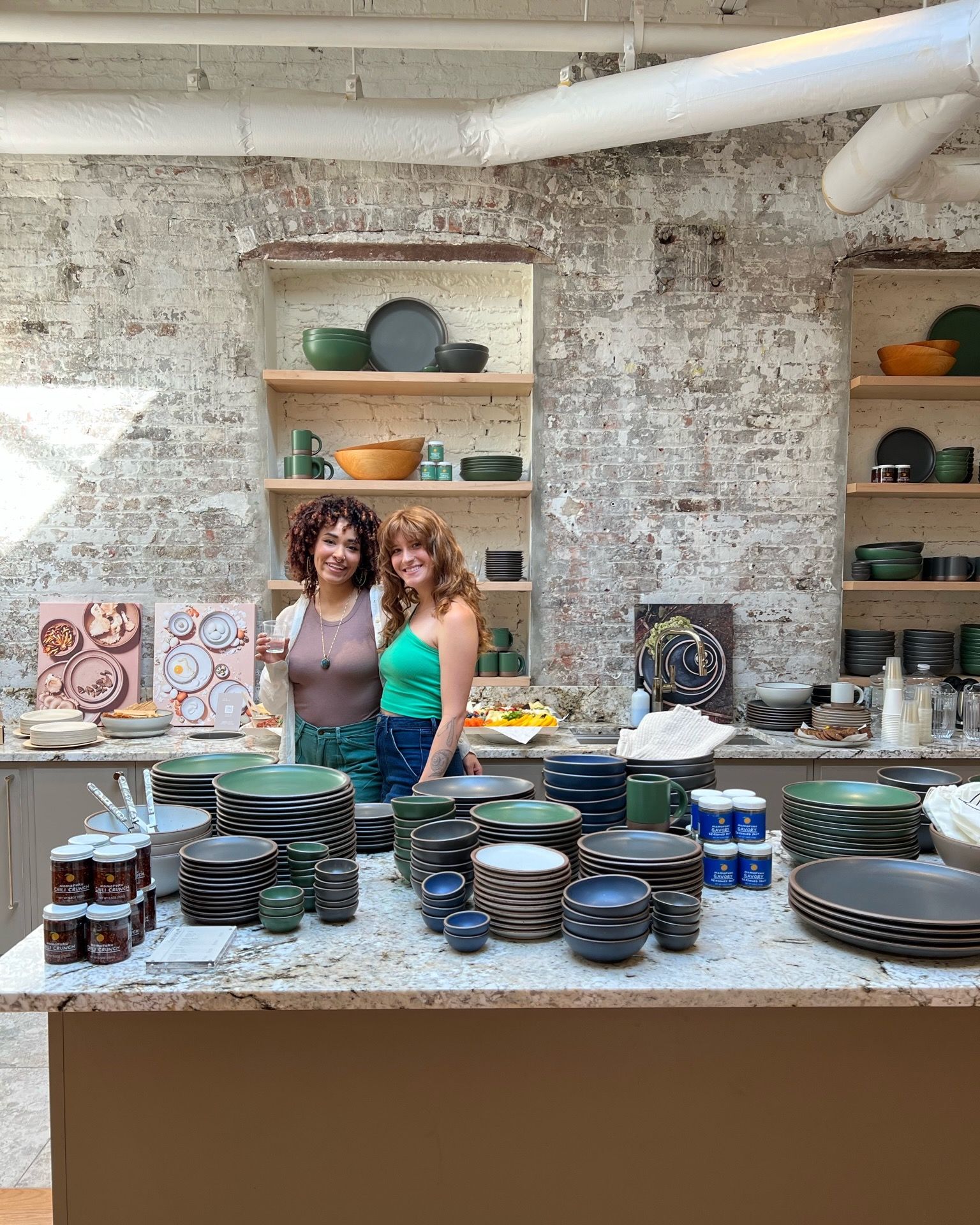 In a retail setting, 2 people are smiling at the camera surrounded by arranged stacks of ceramic bowls and plates merchandised for shopping.
