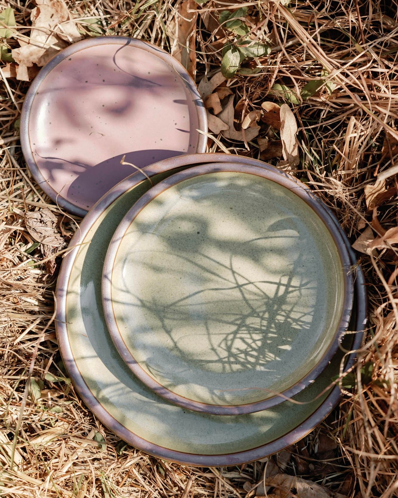 3 Plates of varying sizes in reflective dappled grey-green and dusty mauve pink artfully arranged in dried grass with shadows.