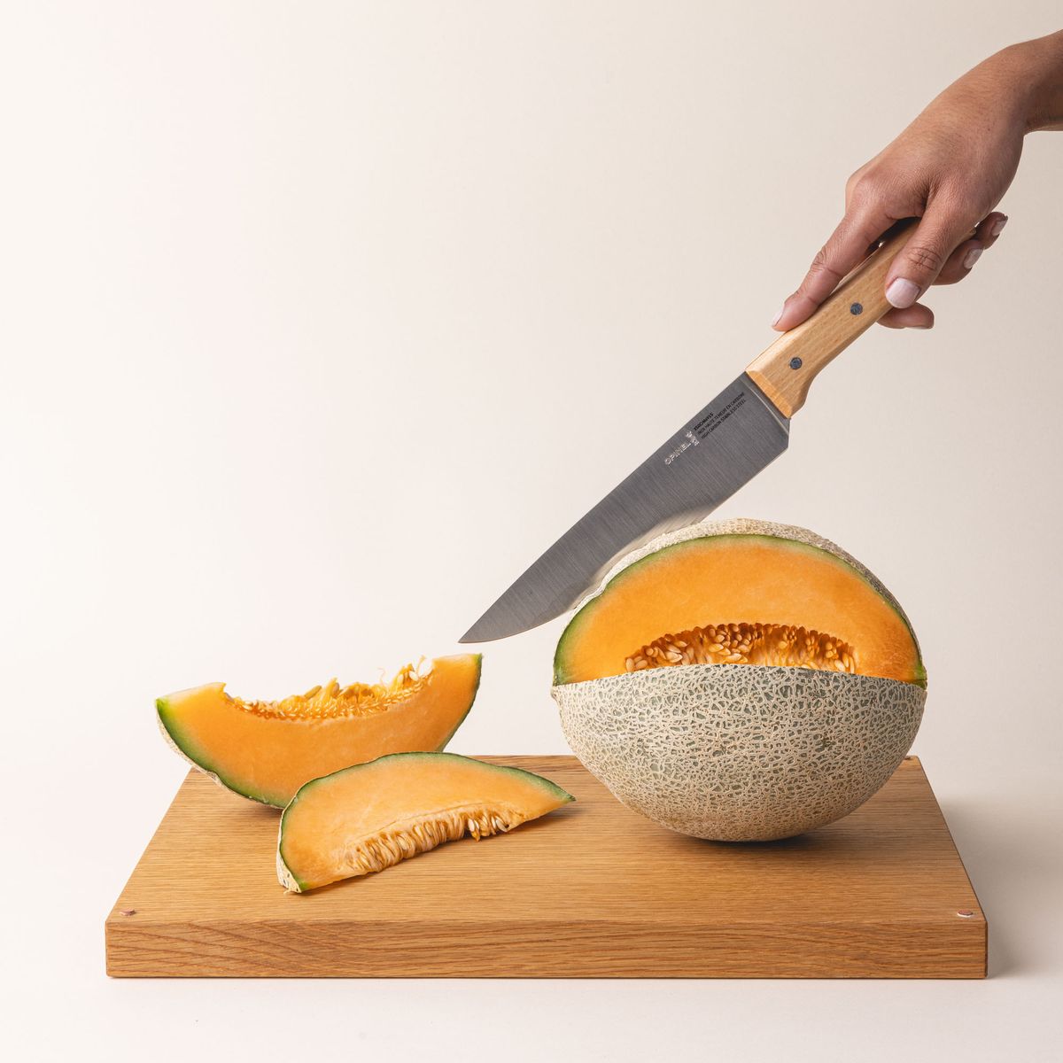 A hand holds a chef's knife with a steel blade and a light wooden handle and is cutting open a cantaloupe on a wooden cutting board.