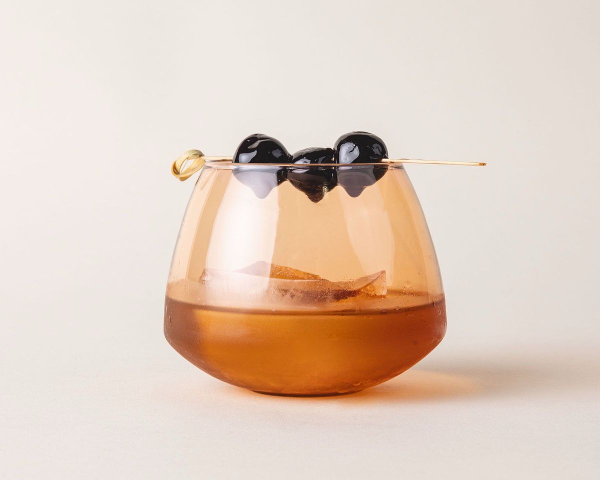 A cocktail with a blueberry garnish in a light orange glass whiskey snifter with a rounded base