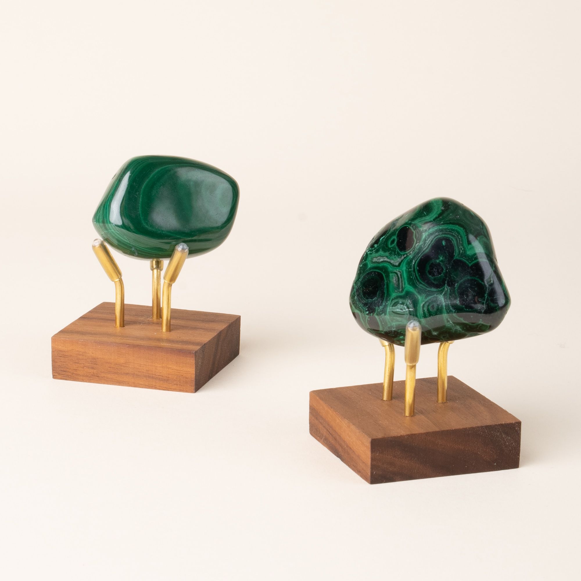 Two polished green malachite stones displayed upright on small square wooden bases with gold supports, set against a neutral background.