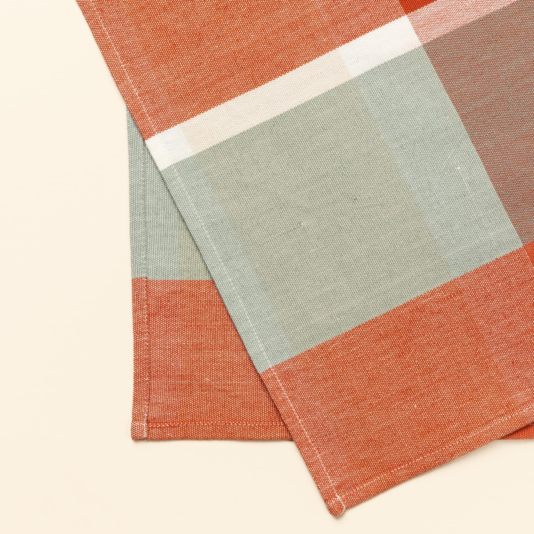 A close up of a corner of a gingham table runner featuring colors of sage green, bold orange, and cream.