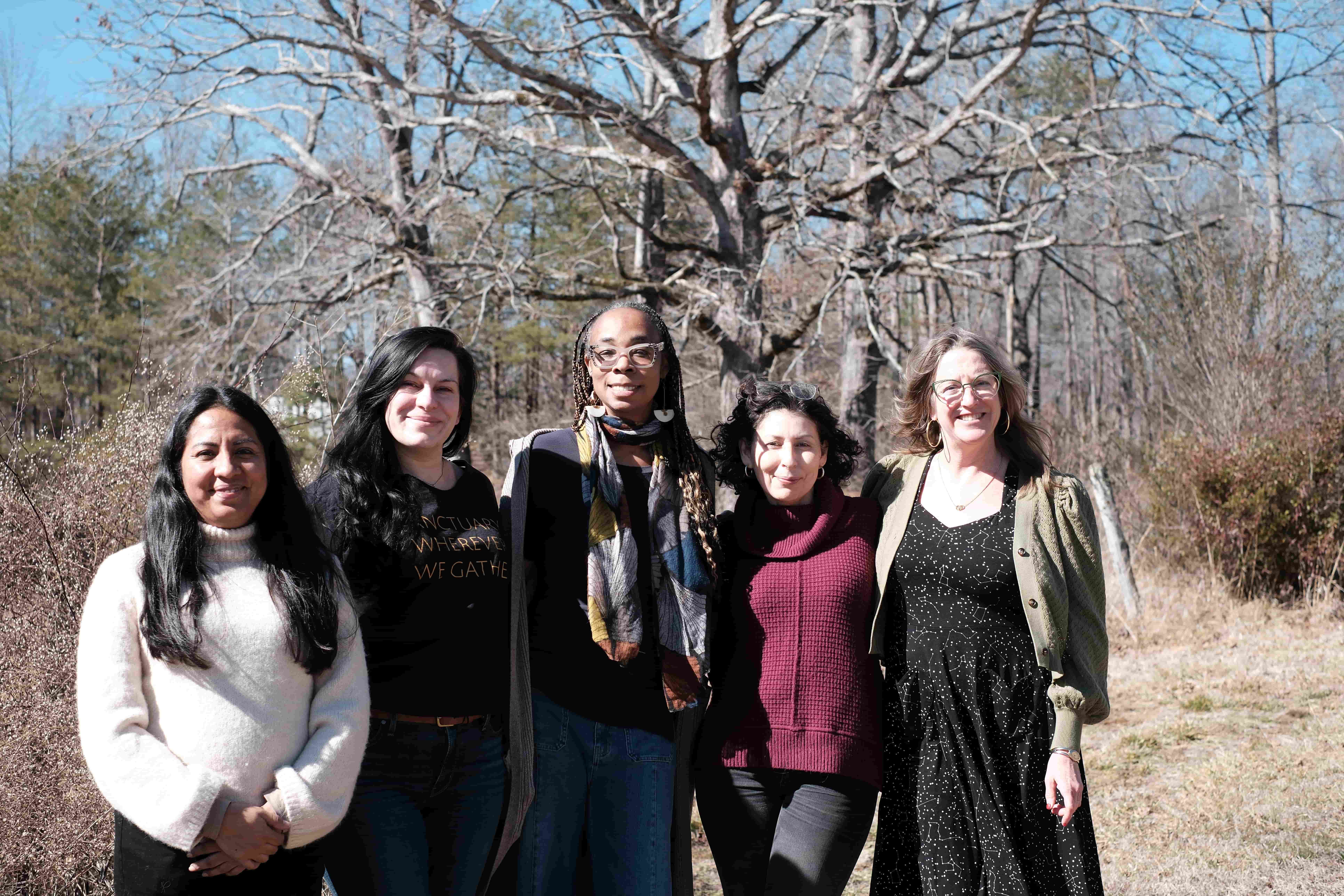 Five women smile with their arms around each other outdoors.