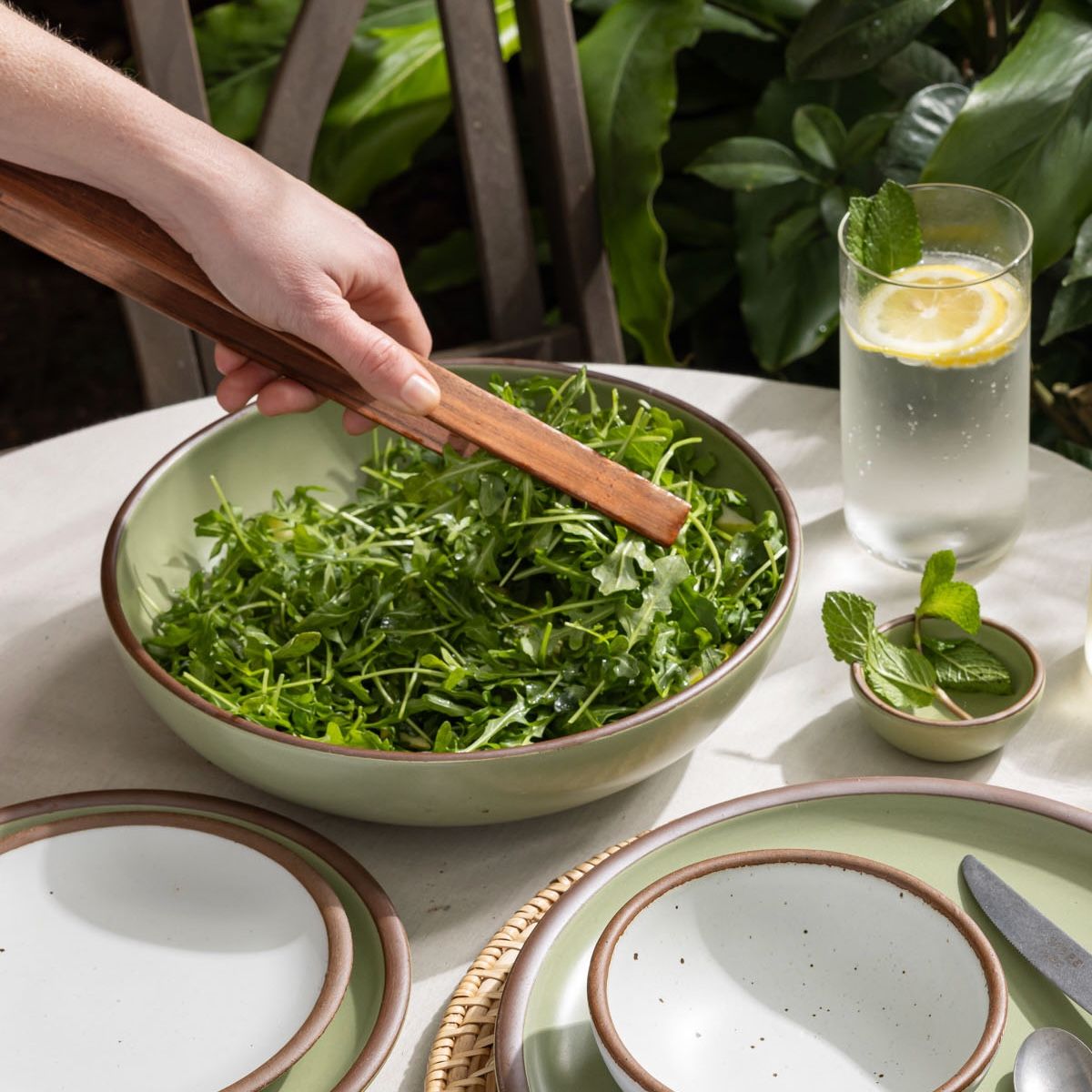 On an outdoor table, a hand holds simple and minimal wood rectangular tongs reaching for arugula in a large serving bowl