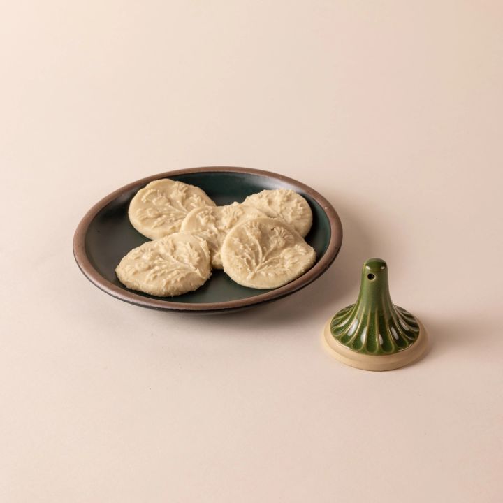 A green ceramic cookie stamp with a cream colored base sits next to a ceramic plate of cookies stamped with a thistle design.