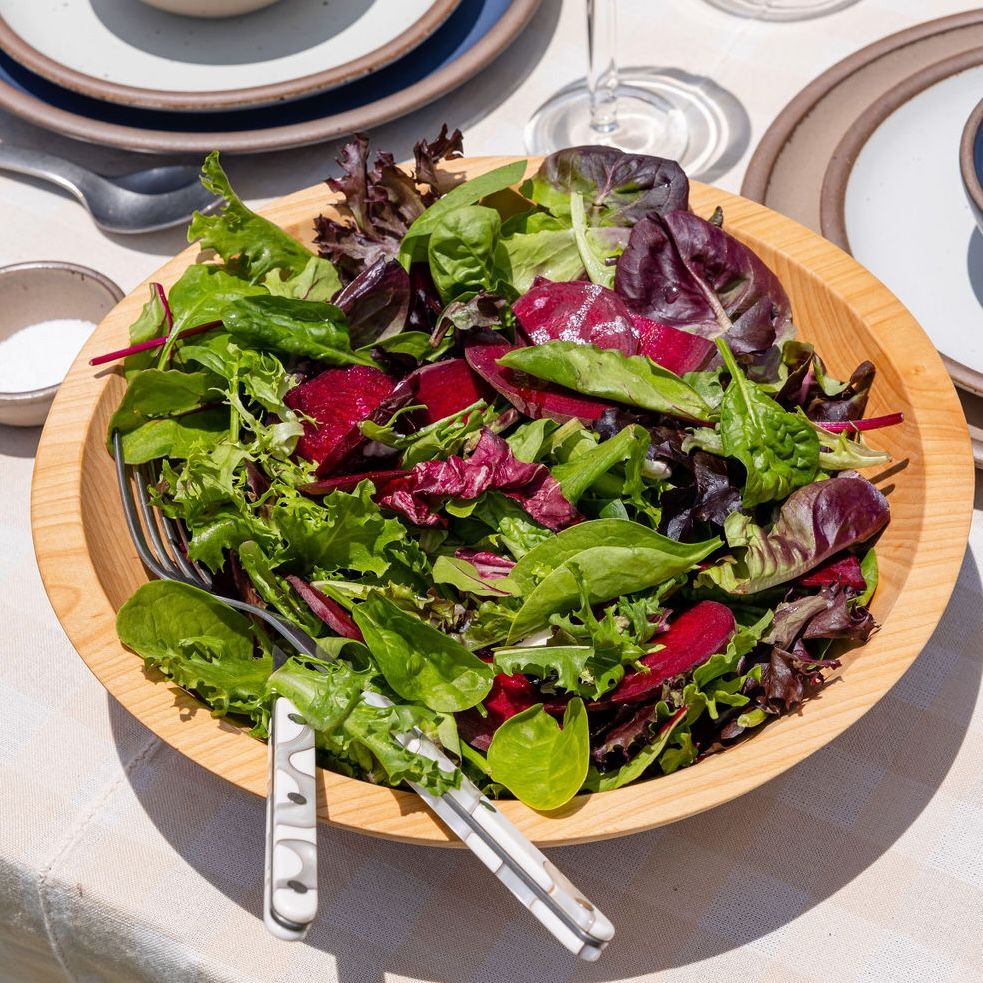A large wooden bowl filled with salad and a serving fork and spoon on top. The bowl is sitting on a table outside.