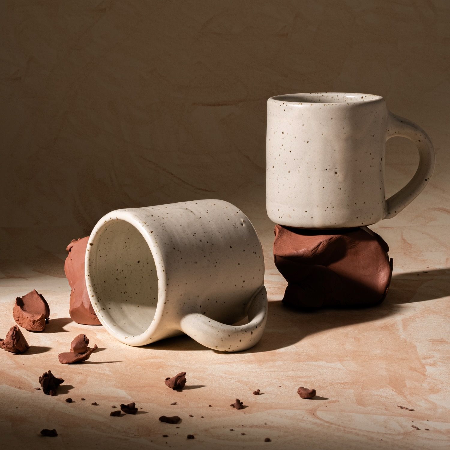 Two medium sized ceramic mugs with handles in a warm, tan-toned, off-white color featuring iron speckles, styled with raw clay