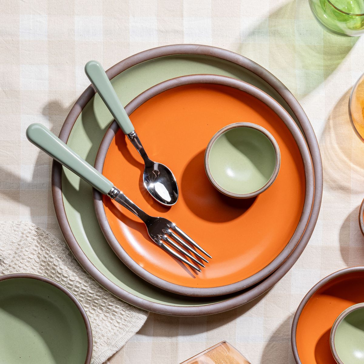 A place setting featuring 2 ceramic plates in orange and sage green with a matching green handled fork and spoon.