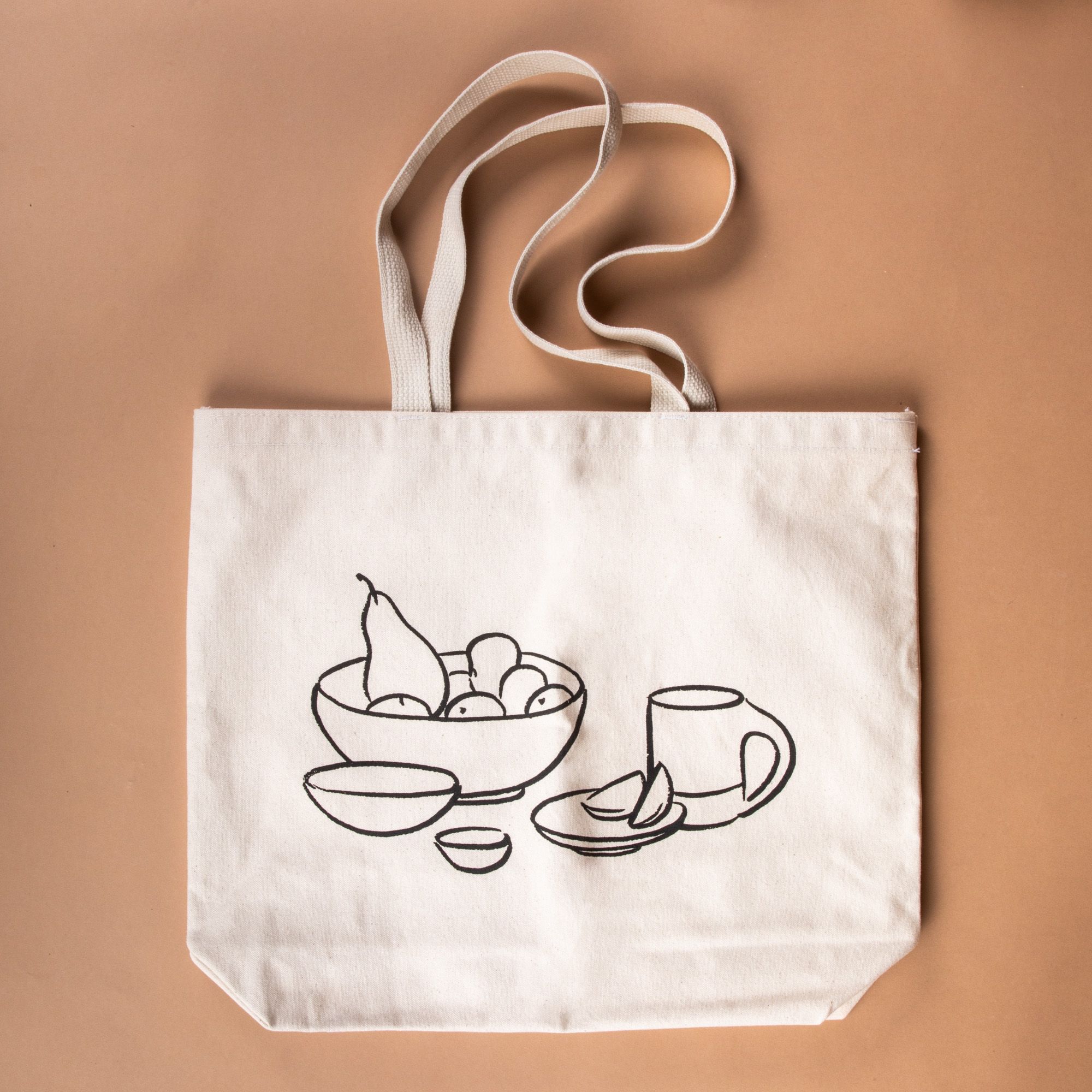 Person holding a cream canvas tote on their shoulder with their hand holding onto the handle - the tote has a charcoal illustration on the front of a bowl with fruit, little bowls, plate, and a mug. Underneath, it reads 'East Fork is a vessel'.