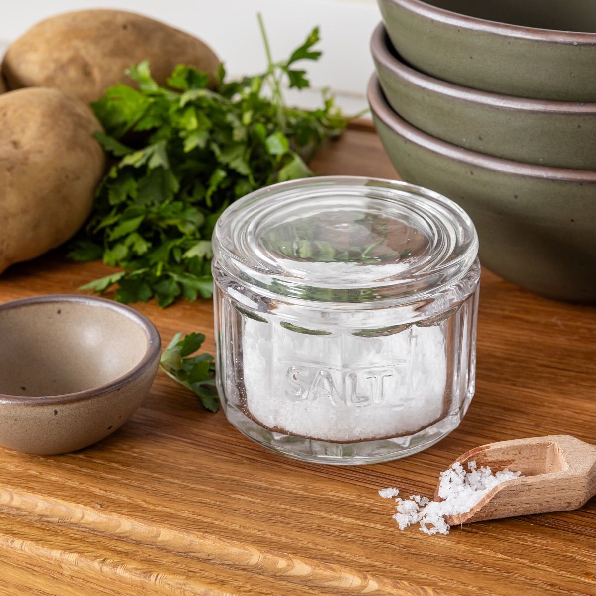 On a shelf is a round glass clear salt canister filled with salt and a small wooden scoop, along with a stack of forest green ceramic bowls.