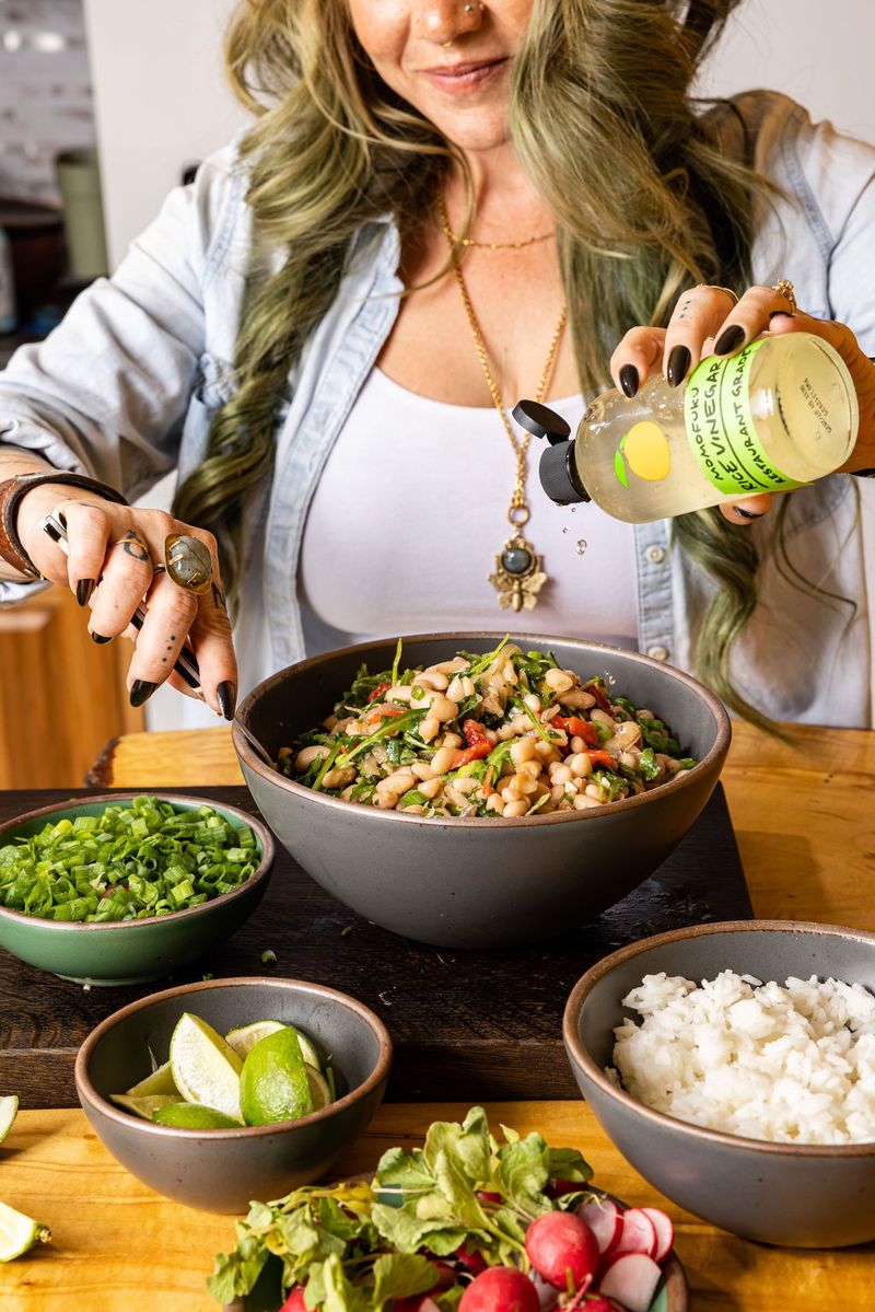 A person pouring rice vinegar into a large grey mixing bowl filled with a bean salad surrounded by other three smaller green and grey bowls filled with scallions, limes, and rice.