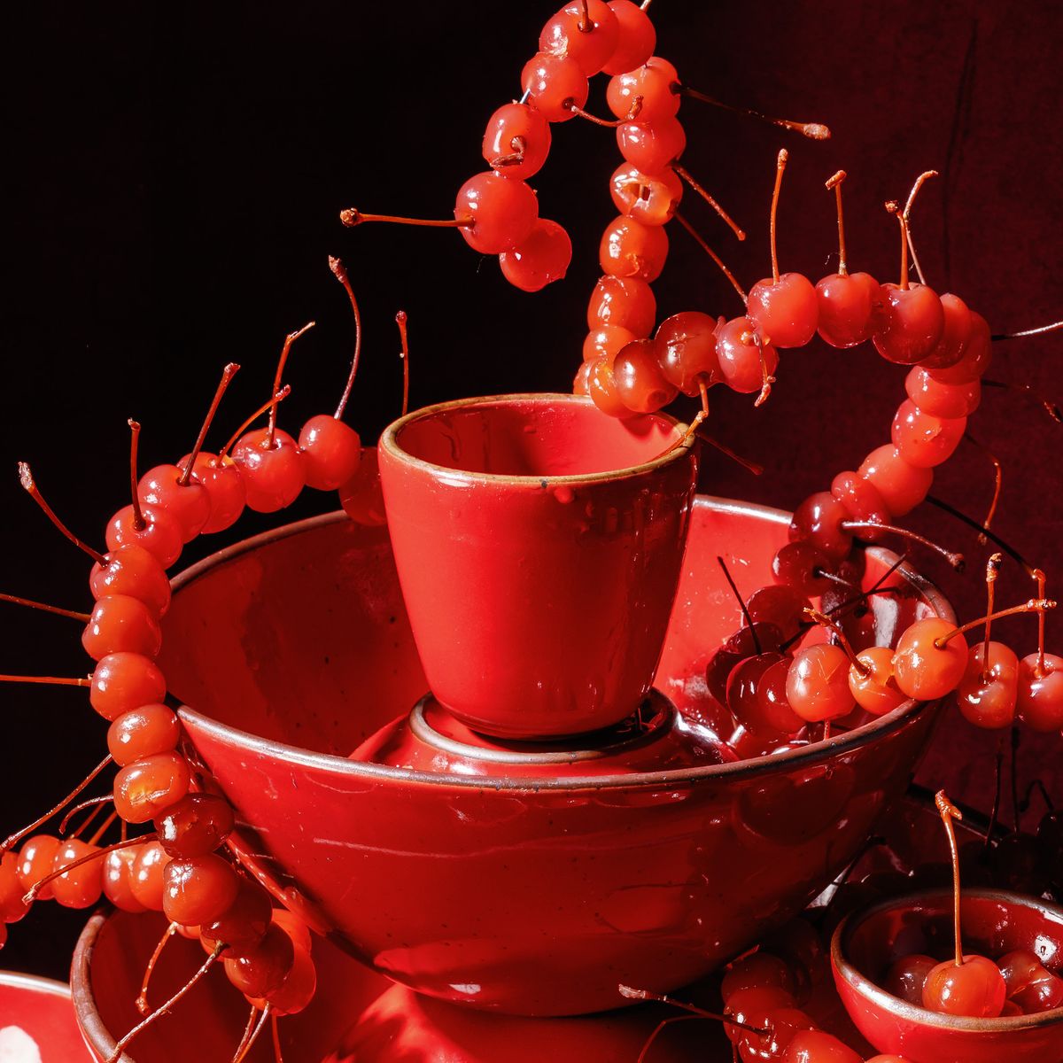 Glossy red ceramic bowls and a tapered cup entwined with strands of bright red cherries against a deep red background.
