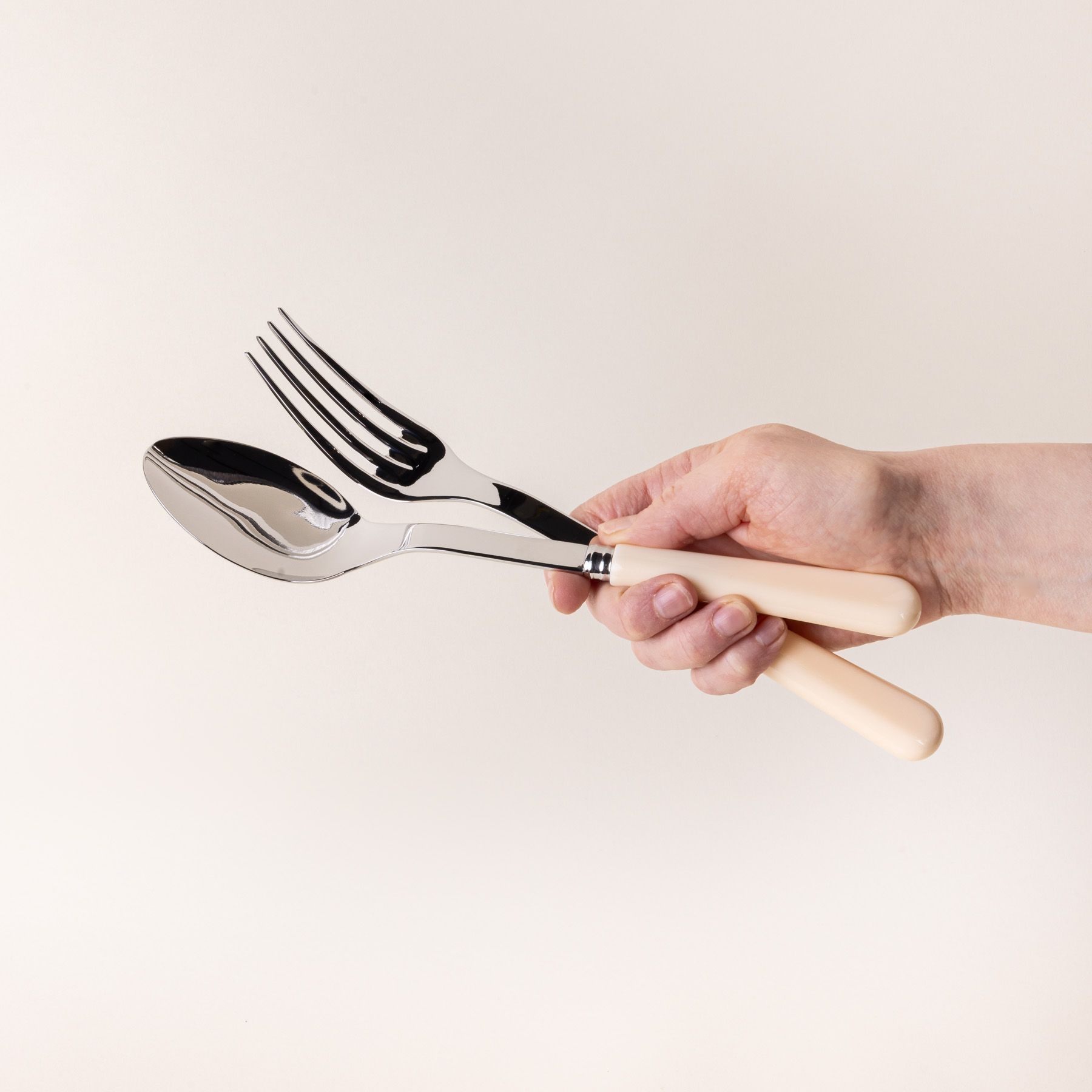A hand holds a serving size spoon and fork in a shiny steel with a matte cream handle