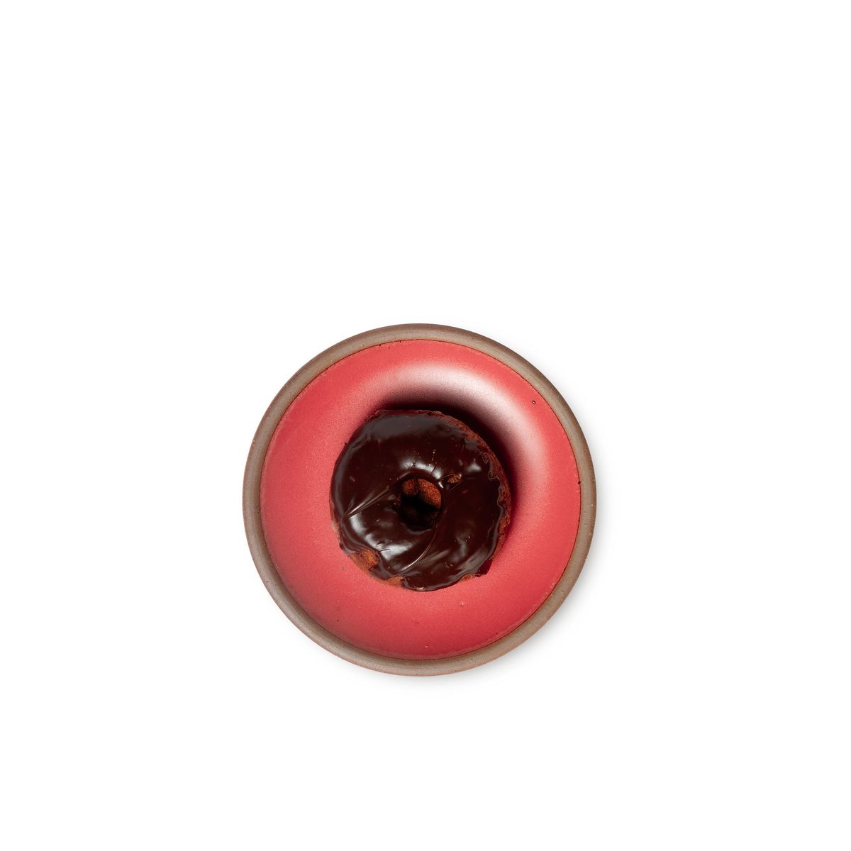 A chocolate donut sits on a dessert sized ceramic plate in a bold red color featuring iron speckles and an unglazed rim