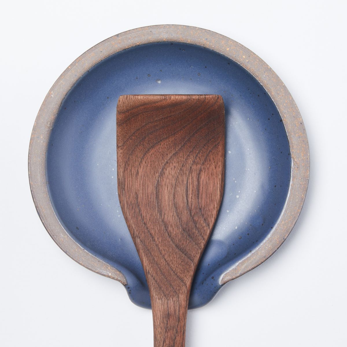 A wooden spatula sits on a circular ceramic spoon rest in a muted navy color featuring iron speckles and unglazed ridge rim