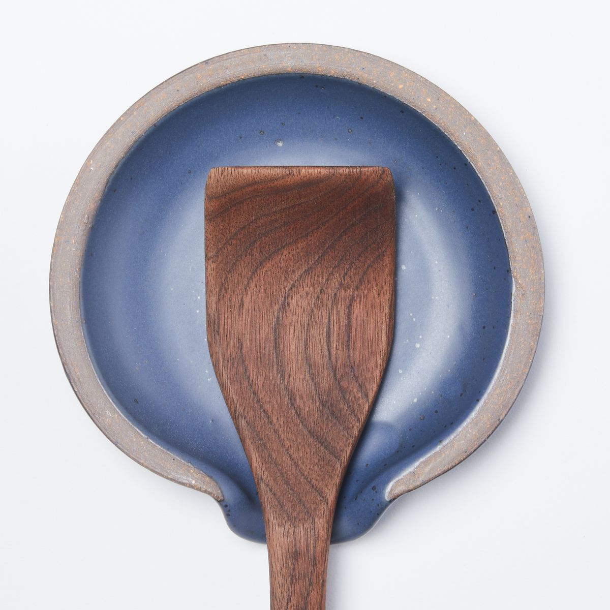 A wooden spatula sits on a circular ceramic spoon rest in a muted navy color featuring iron speckles and unglazed ridge rim