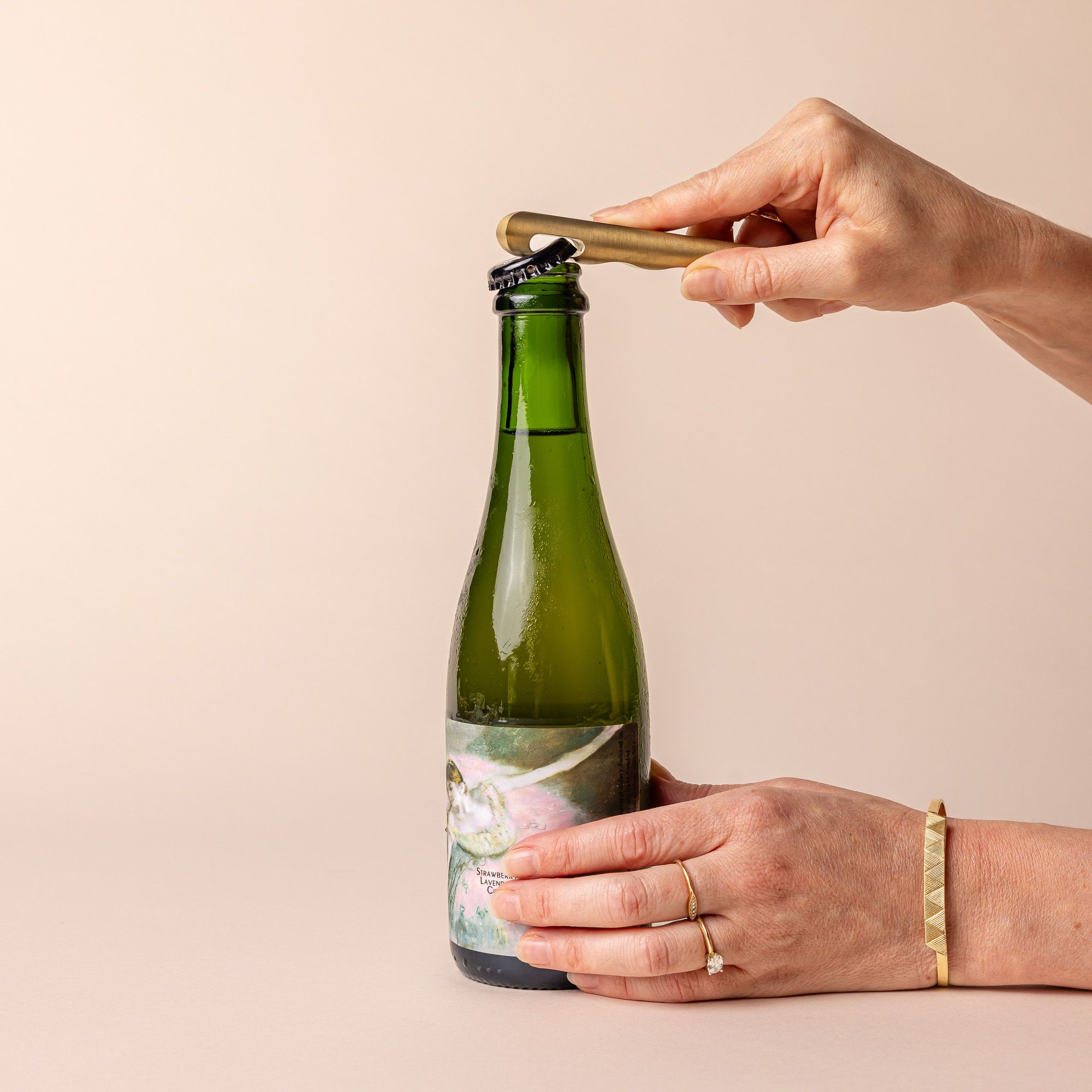 Hands holding a green bottle and a sophisticated brass bottle opener to open it.