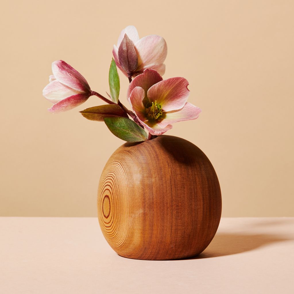 Three pink flowers with green leaves emerge from the top of a wooden vase that is shaped like an orb